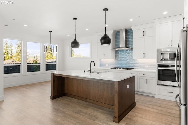 kitchen with appliances with stainless steel finishes, a kitchen island with sink, sink, white cabinetry, and wall chimney range hood