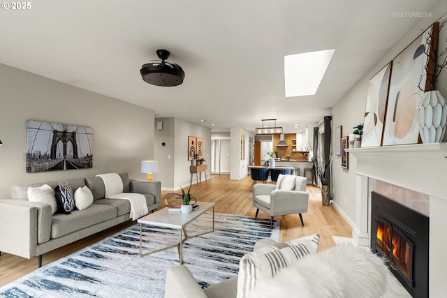 living room with baseboards, a skylight, light wood-style floors, a fireplace, and ceiling fan