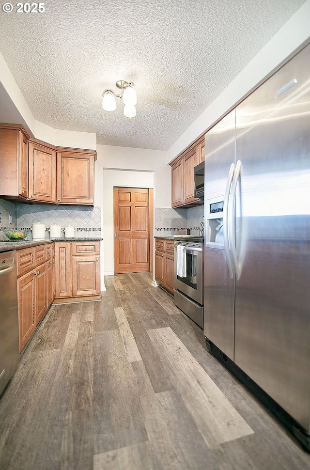 kitchen with a textured ceiling, stainless steel appliances, wood finished floors, decorative backsplash, and brown cabinets