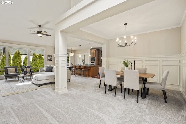 dining room with a decorative wall, light carpet, ceiling fan with notable chandelier, wainscoting, and crown molding