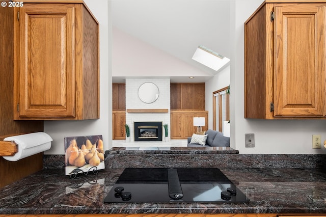 kitchen with brown cabinets, lofted ceiling with skylight, a brick fireplace, dark stone counters, and black electric cooktop