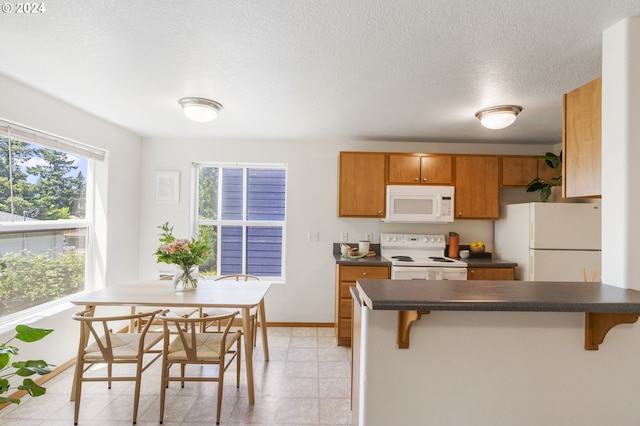 kitchen with dark countertops, brown cabinetry, white appliances, a peninsula, and a kitchen bar