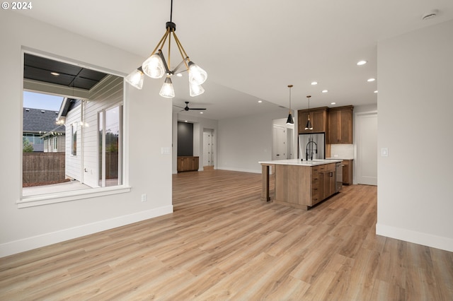 kitchen with light wood-type flooring, ceiling fan with notable chandelier, stainless steel appliances, pendant lighting, and an island with sink