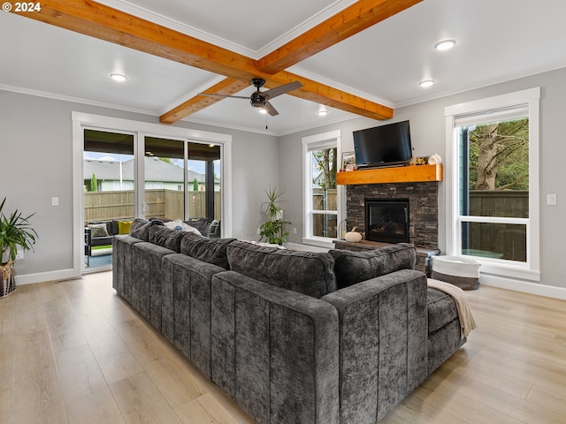 living room with ceiling fan, beamed ceiling, ornamental molding, a stone fireplace, and light wood-type flooring