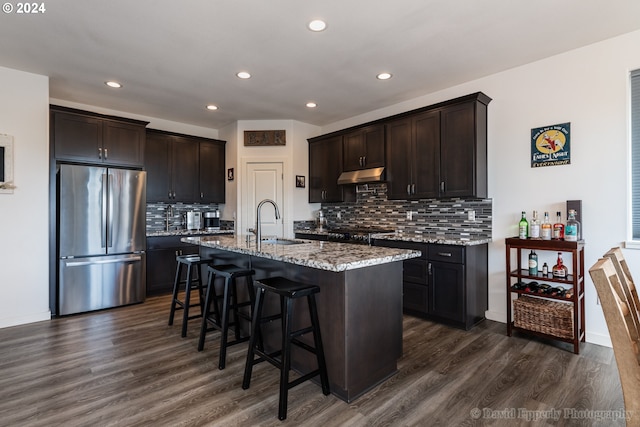 kitchen featuring sink, dark wood-type flooring, stainless steel fridge, tasteful backsplash, and a center island with sink