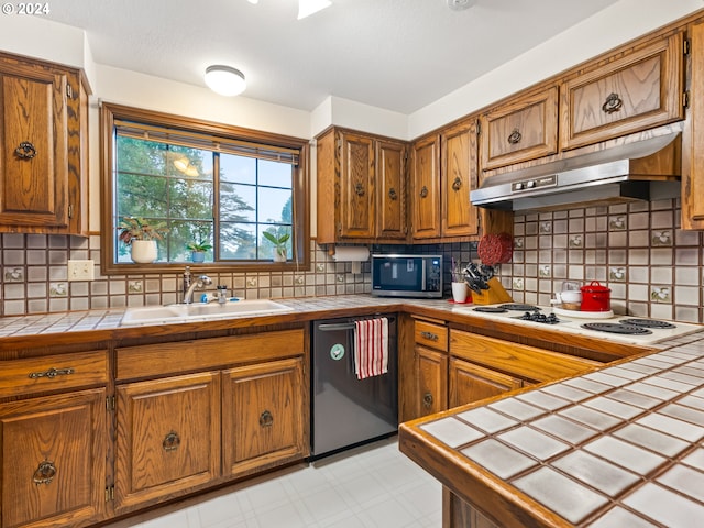 kitchen with tile countertops, dishwashing machine, stainless steel microwave, under cabinet range hood, and a sink