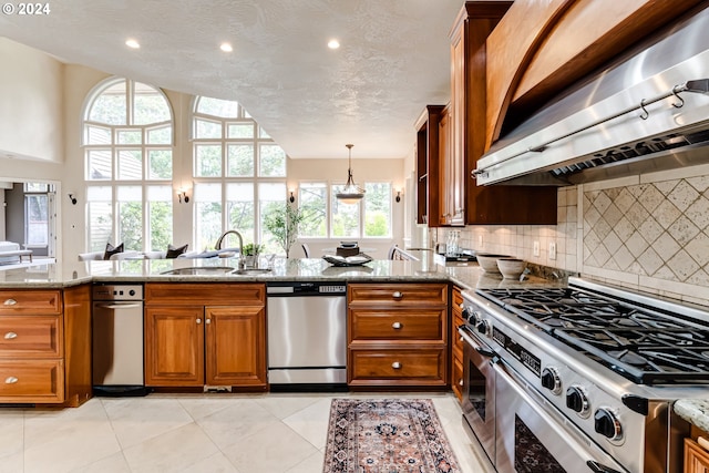 kitchen featuring appliances with stainless steel finishes, brown cabinetry, a sink, wall chimney range hood, and light stone countertops