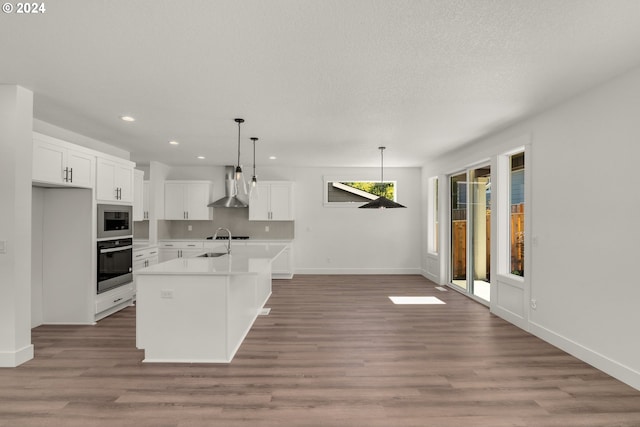 kitchen with white cabinetry, wall chimney range hood, oven, a center island with sink, and built in microwave