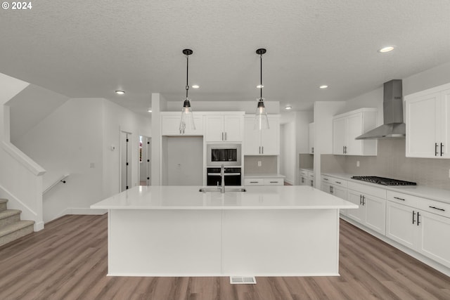 kitchen featuring decorative light fixtures, wall chimney range hood, stainless steel gas cooktop, an island with sink, and white cabinets