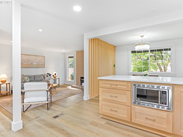 kitchen with stainless steel microwave, a brick fireplace, light brown cabinetry, decorative light fixtures, and light hardwood / wood-style floors