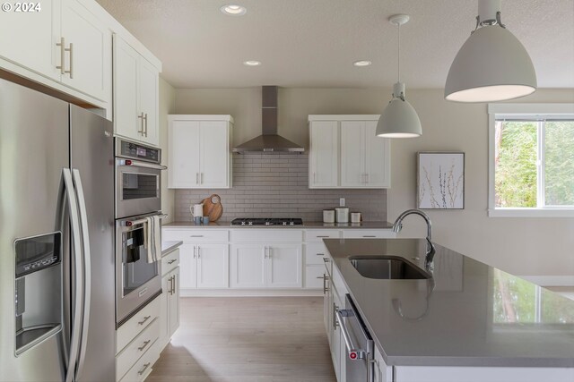 kitchen with appliances with stainless steel finishes, hanging light fixtures, white cabinets, wall chimney exhaust hood, and a center island with sink