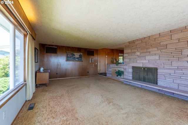 unfurnished living room featuring light colored carpet, a textured ceiling, a stone fireplace, and wooden walls