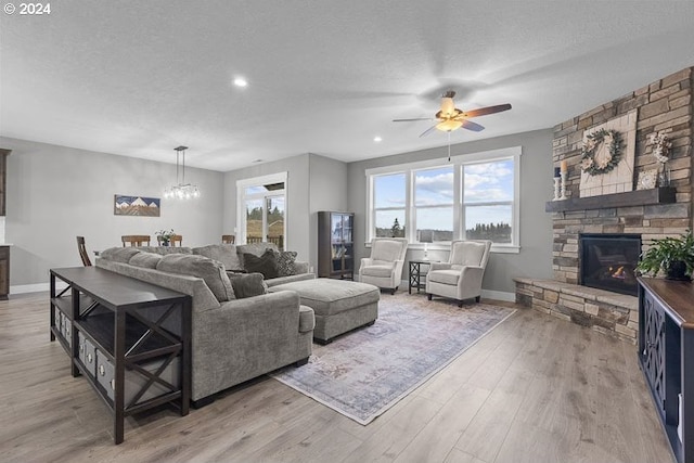 living room featuring a textured ceiling, a stone fireplace, light hardwood / wood-style floors, and ceiling fan with notable chandelier