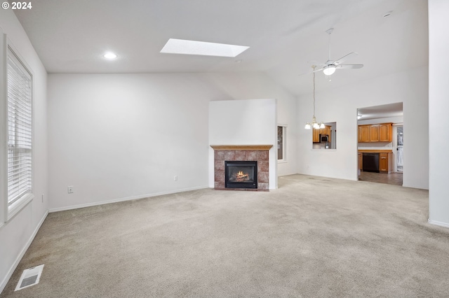 unfurnished living room featuring a tile fireplace, light carpet, ceiling fan with notable chandelier, and vaulted ceiling with skylight