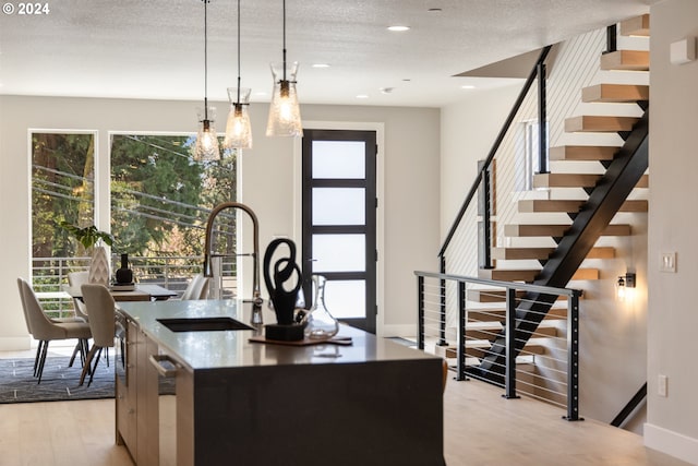 kitchen with decorative light fixtures, a textured ceiling, an island with sink, sink, and light wood-type flooring