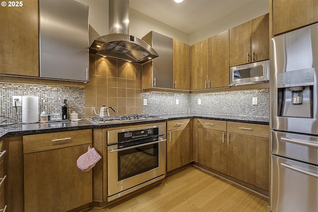 kitchen featuring appliances with stainless steel finishes, backsplash, exhaust hood, light hardwood / wood-style flooring, and dark stone countertops