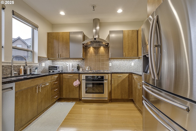 kitchen featuring dark stone counters, sink, light wood-type flooring, island range hood, and stainless steel appliances