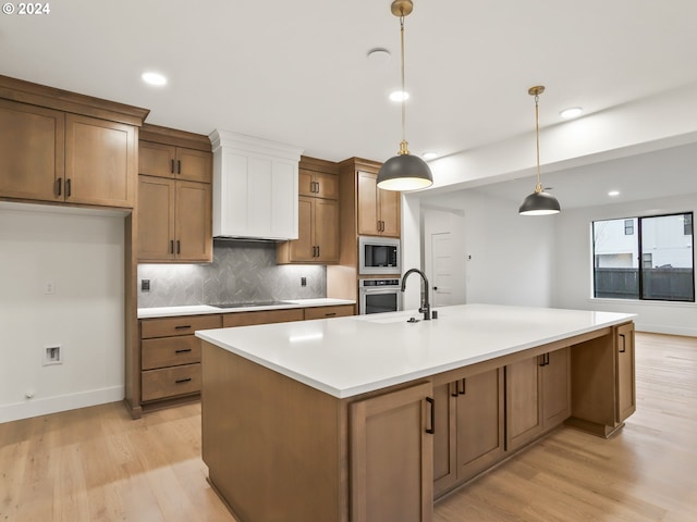 kitchen with light wood-type flooring, tasteful backsplash, stainless steel appliances, a kitchen island with sink, and pendant lighting