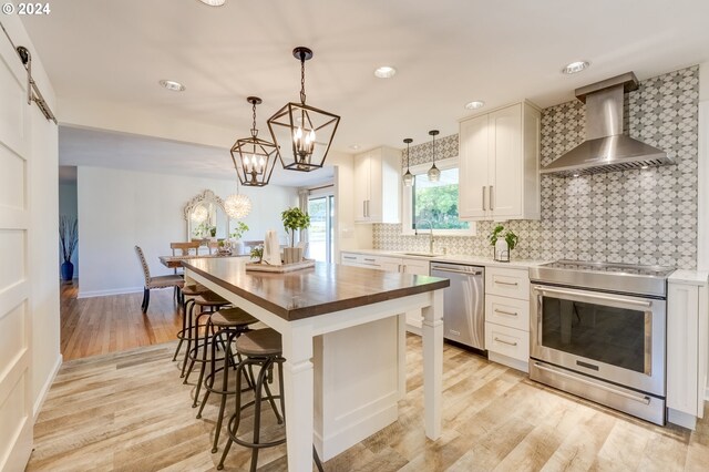 kitchen featuring butcher block countertops, a breakfast bar area, wall chimney exhaust hood, appliances with stainless steel finishes, and white cabinets