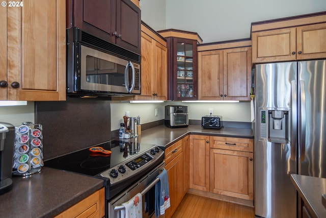 kitchen featuring light wood finished floors, glass insert cabinets, appliances with stainless steel finishes, dark countertops, and brown cabinets