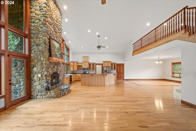 unfurnished living room with a high ceiling, a stone fireplace, ceiling fan with notable chandelier, and light hardwood / wood-style flooring