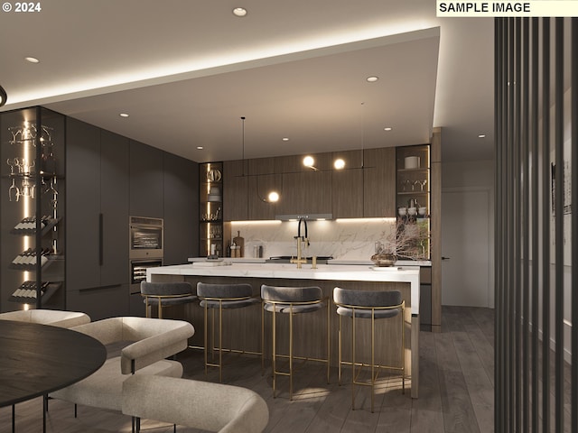 kitchen with hanging light fixtures, tasteful backsplash, a kitchen island with sink, dark wood-type flooring, and a kitchen breakfast bar