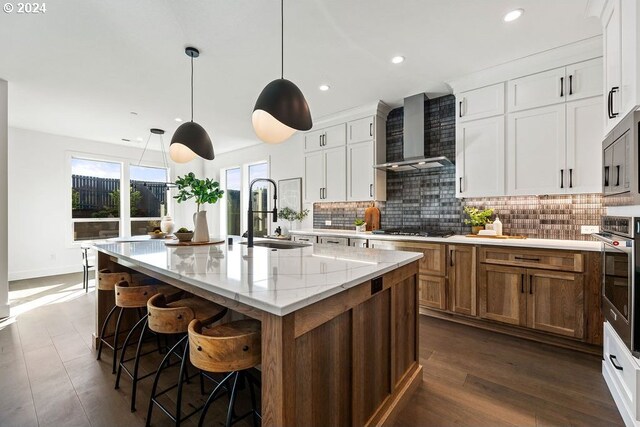 kitchen with wall chimney range hood, sink, appliances with stainless steel finishes, a kitchen island with sink, and light stone counters