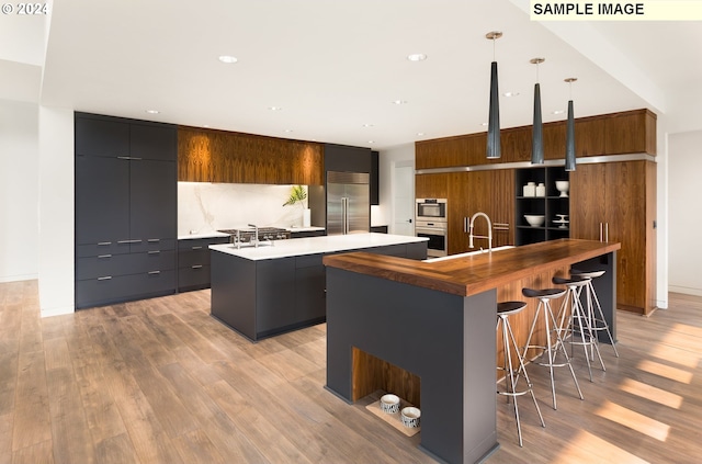 kitchen featuring backsplash, appliances with stainless steel finishes, hanging light fixtures, an island with sink, and light hardwood / wood-style flooring