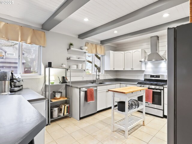 kitchen with wall chimney exhaust hood, stainless steel appliances, light tile patterned floors, beamed ceiling, and white cabinetry