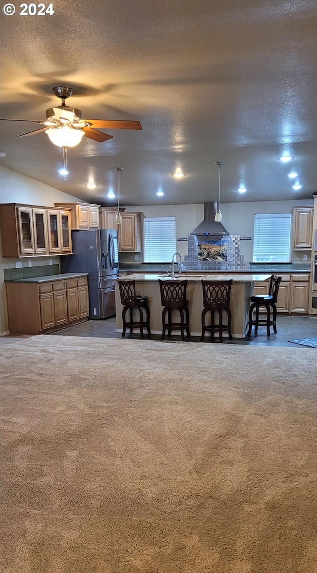 kitchen with hanging light fixtures, wall chimney range hood, stainless steel refrigerator with ice dispenser, a textured ceiling, and a kitchen bar