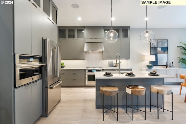 kitchen featuring stainless steel appliances, pendant lighting, wall chimney exhaust hood, light hardwood / wood-style flooring, and a kitchen island with sink