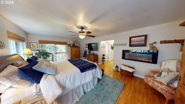 bedroom featuring access to outside, a glass covered fireplace, wood finished floors, baseboards, and ceiling fan