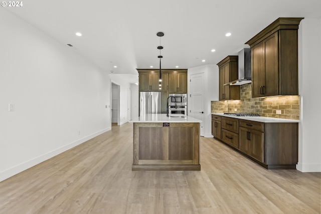 kitchen with wall chimney range hood, an island with sink, stainless steel appliances, and light hardwood / wood-style flooring