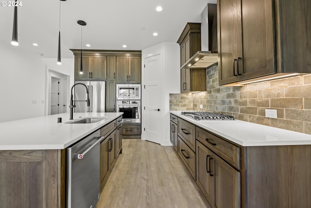 kitchen featuring wall chimney range hood, light hardwood / wood-style floors, appliances with stainless steel finishes, an island with sink, and sink