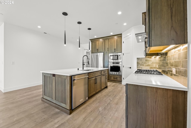 kitchen featuring stainless steel appliances, a center island with sink, light wood-type flooring, hanging light fixtures, and sink