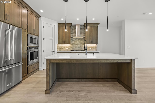 kitchen featuring light hardwood / wood-style floors, appliances with stainless steel finishes, an island with sink, wall chimney exhaust hood, and tasteful backsplash