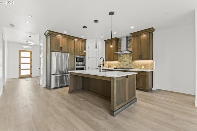 kitchen with wall chimney range hood, stainless steel appliances, light wood-type flooring, and a kitchen island with sink