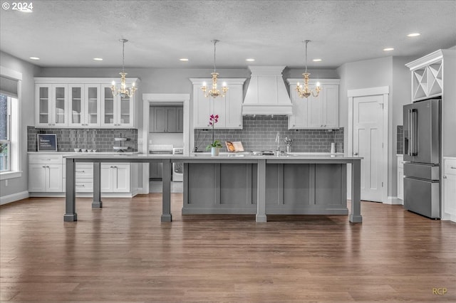 kitchen with white cabinetry, custom exhaust hood, hanging light fixtures, high end fridge, and a center island with sink