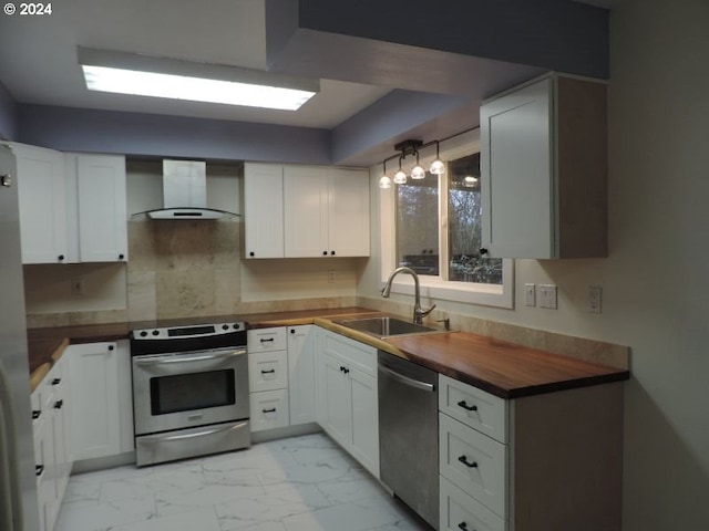 kitchen featuring wood counters, appliances with stainless steel finishes, wall chimney exhaust hood, sink, and white cabinetry