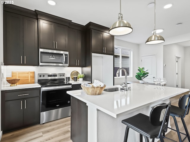 kitchen with pendant lighting, light countertops, appliances with stainless steel finishes, a kitchen island with sink, and a sink