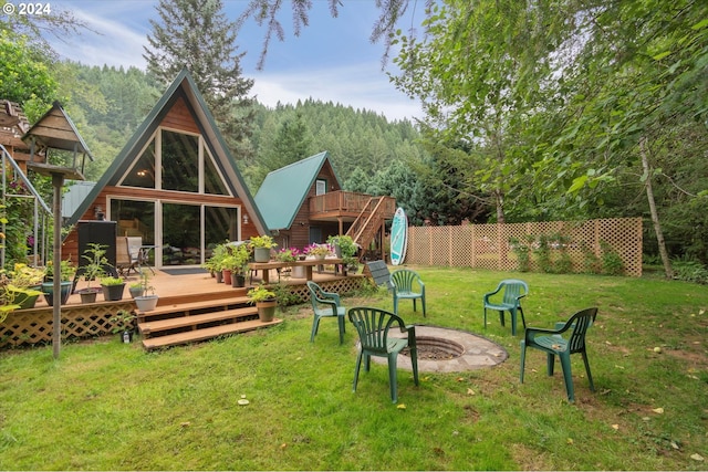 rear view of property featuring a lawn, a wooden deck, an outdoor fire pit, fence, and a forest view