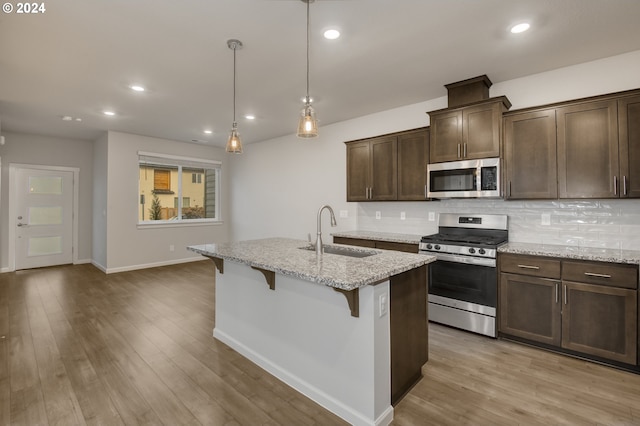 kitchen featuring an island with sink, stainless steel appliances, decorative light fixtures, light stone countertops, and sink