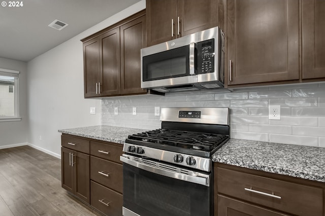 kitchen with stainless steel appliances, tasteful backsplash, dark brown cabinets, light hardwood / wood-style flooring, and light stone counters