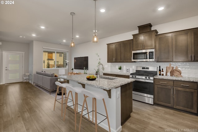 kitchen featuring stainless steel appliances, a kitchen island with sink, decorative light fixtures, light stone counters, and sink