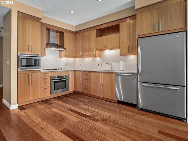 kitchen with backsplash, stainless steel appliances, wall chimney range hood, and light wood-type flooring