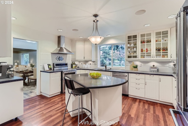 kitchen with dark hardwood / wood-style flooring, appliances with stainless steel finishes, white cabinetry, wall chimney exhaust hood, and decorative light fixtures