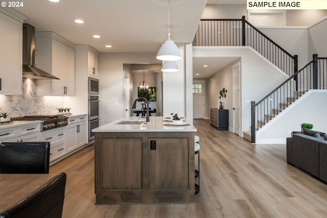 kitchen with light wood-type flooring, appliances with stainless steel finishes, wall chimney range hood, sink, and an island with sink