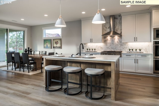 kitchen with wall chimney exhaust hood, pendant lighting, light hardwood / wood-style flooring, backsplash, and a center island with sink