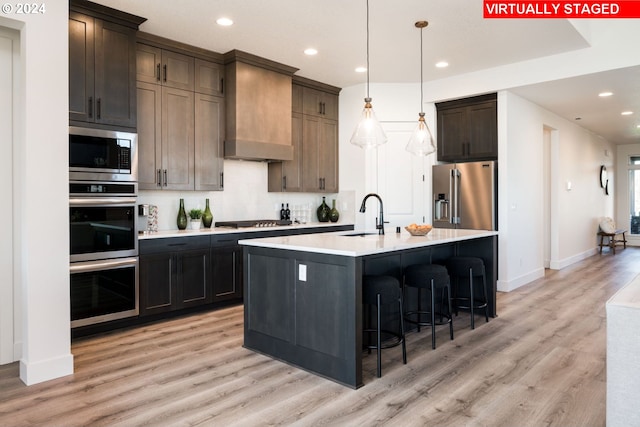 kitchen with custom range hood, stainless steel appliances, a kitchen island with sink, sink, and decorative light fixtures