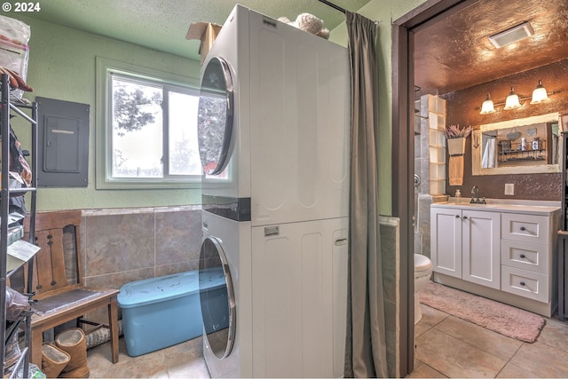 clothes washing area featuring stacked washing maching and dryer, a textured ceiling, sink, light tile patterned floors, and electric panel